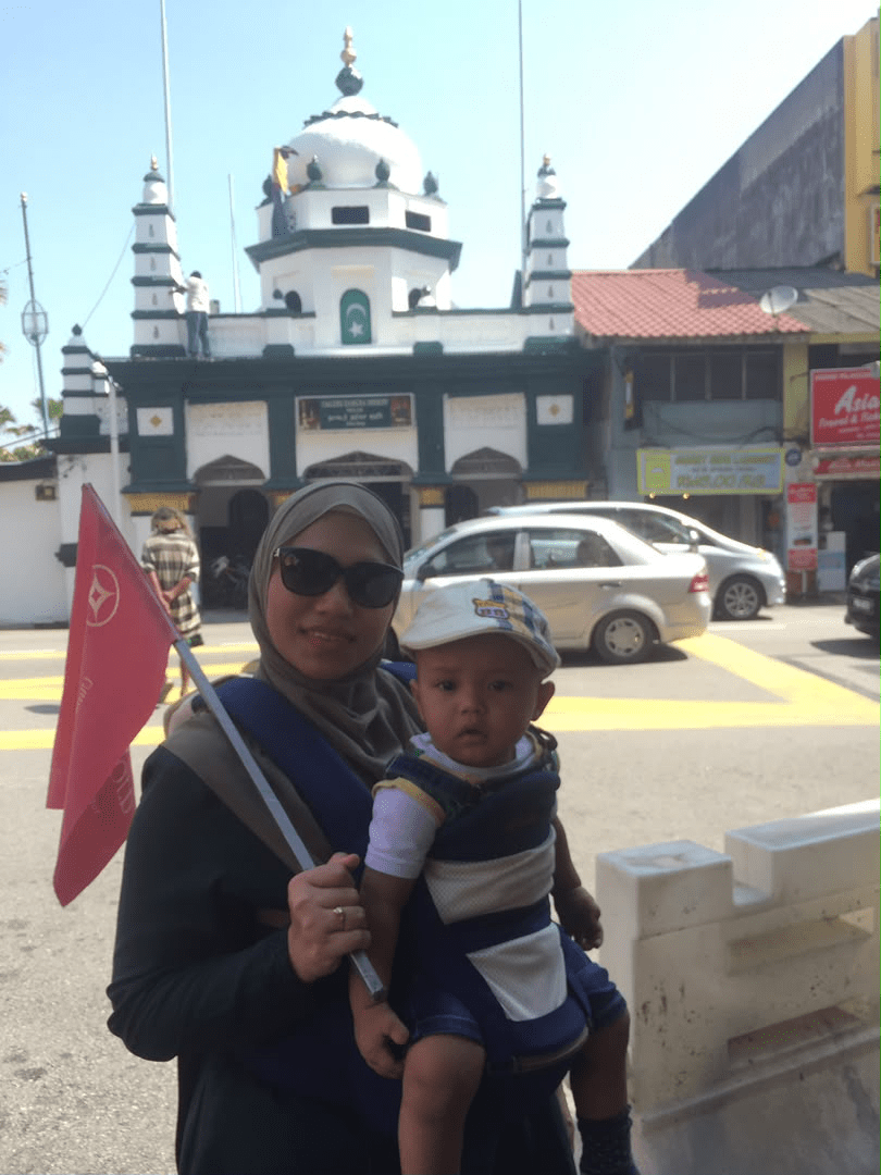 Puan Syamimi mengendong anak kecil sambil memegang bendera Public Gold semasa aktiviti promosi di George Town
