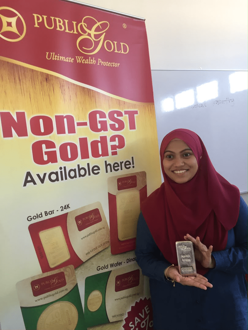 Puan Syamimi memegang jongkong perak Public Gold 1 Kilogram di samping bunting Non-GST Gold