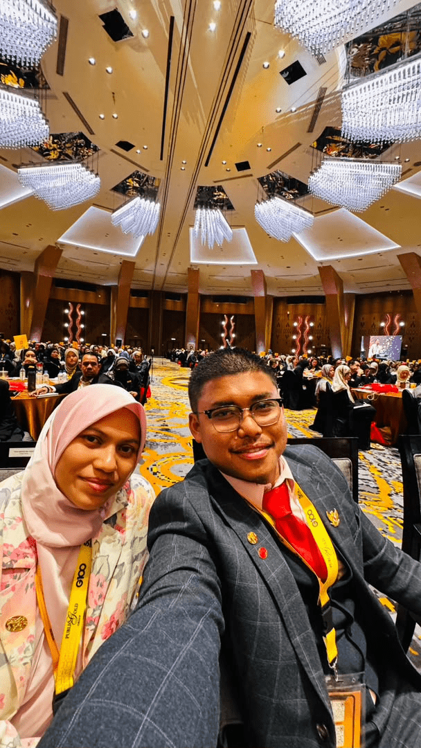 Syarif Jamil (SJ) dan Puan Syamimi di dewan besar berkilauan lampu candelier bersama ratusan barisan dealer empayar di belakang mereka